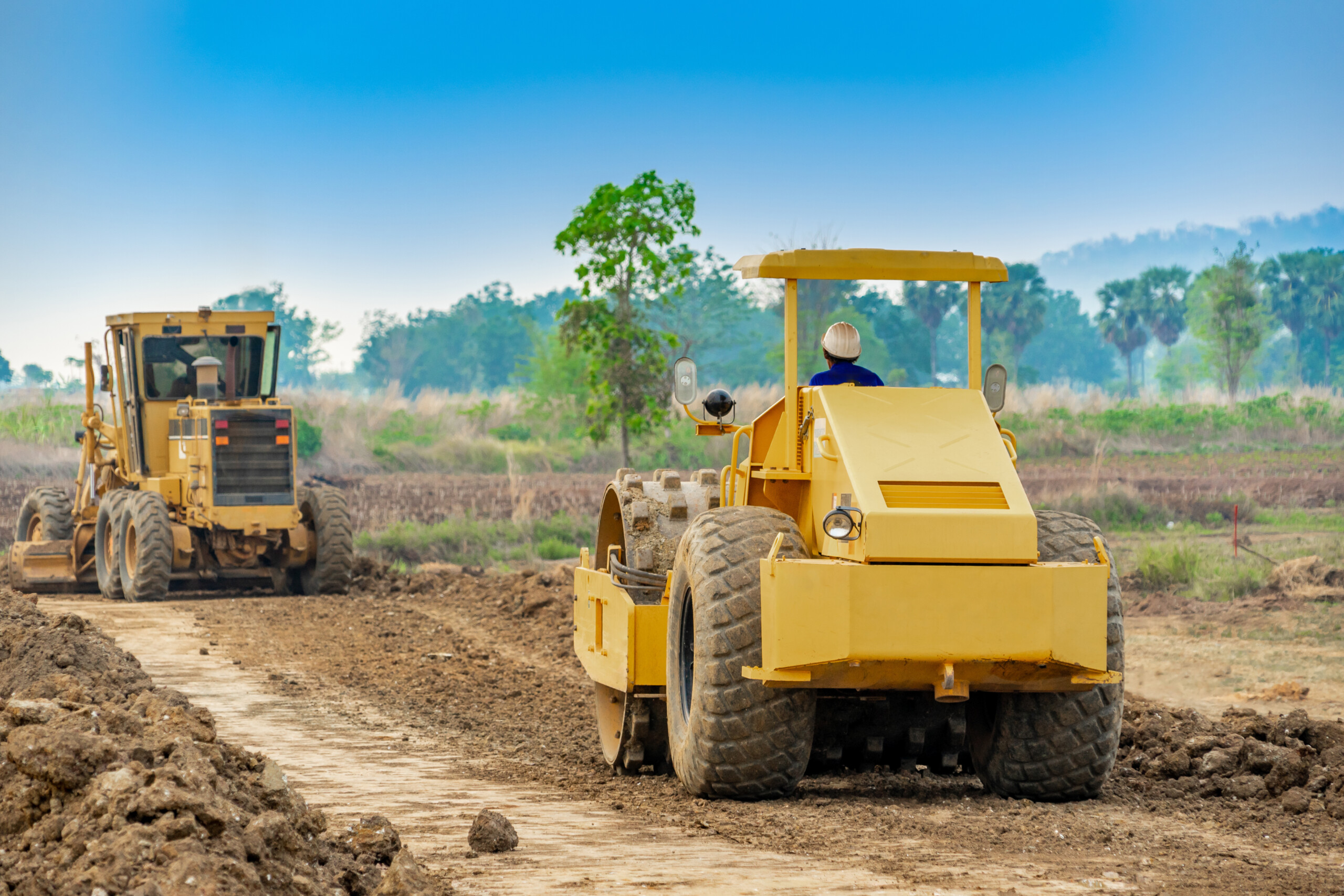 The selective focus vibratory sheepsfoot roller compactor working on construction site and following Motor grader clearing and leveling construction site surface.